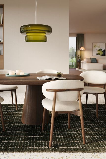 Eethoek met ronde donkerhouten tafel en licht gestoffeerde stoelen, onder een groene hanglamp; op de achtergrond een open leefruimte.