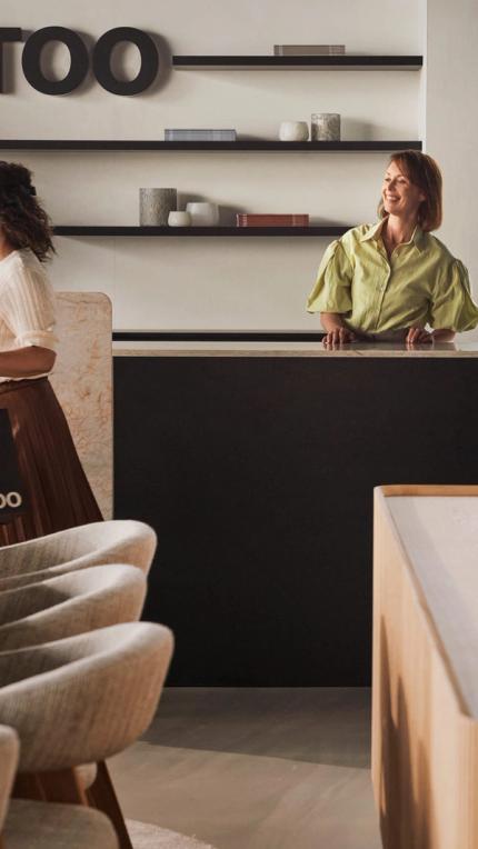 Vrouw in een groene blouse die glimlacht en een marmeren balie aanraakt, omringd door moderne decoratieve objecten in een showroom