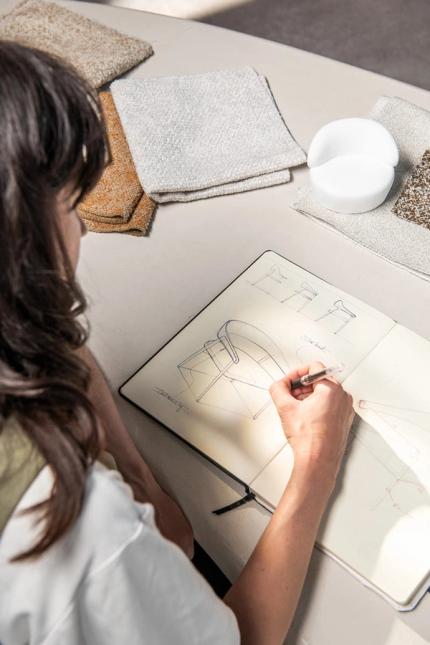 Persoon maakt een ontwerpschets in een open schetsboek op tafel, met stofstalen en een wit modelletje ernaast.