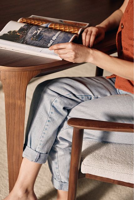 Persoon leest een open magazine aan een houten tafel, zittend op een stoel met houten armleuning en lichte stoffen zitting.