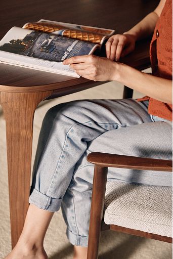 Persoon leest een open magazine aan een houten tafel, zittend op een stoel met houten armleuning en lichte stoffen zitting.