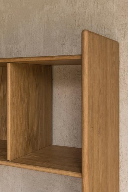 Close-up van de bovenhoek van een houten boekenkast, met afgeronde zijplaat en open vak tegen een ruwe muur.