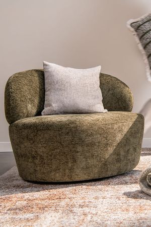 Fauteuil arrondi en tissu vert olive richement texturé, avec un large dossier et un coussin décoratif clair, posé sur un tapis aux tons chauds.