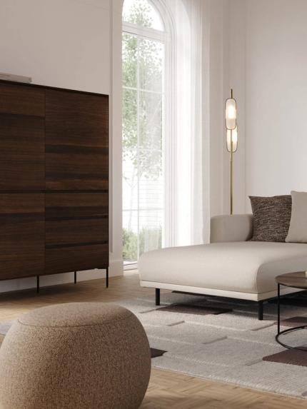 Pouf Basso en tissu beige, placé à côté d’un canapé beige et d’une table basse dans un salon moderne avec un sol en bois et de grandes fenêtres.t houten vloer en grote ramen.