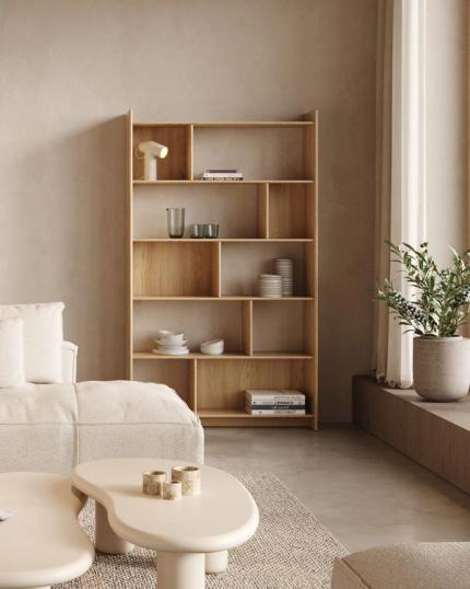 Houten boekenkast met open vakverdeling in een rustige woonkamer, naast een groot raam met gordijnen en witte salontafels op tapijt.