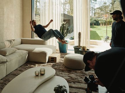 Behind the scenes van een fotoshoot: een man springt in de zetel en wordt in het midden van de sprong zwevend gecapteerd in een woonkamer, terwijl crewleden en opnameapparatuur zichtbaar zijn en de setting van de productie onthullen.
