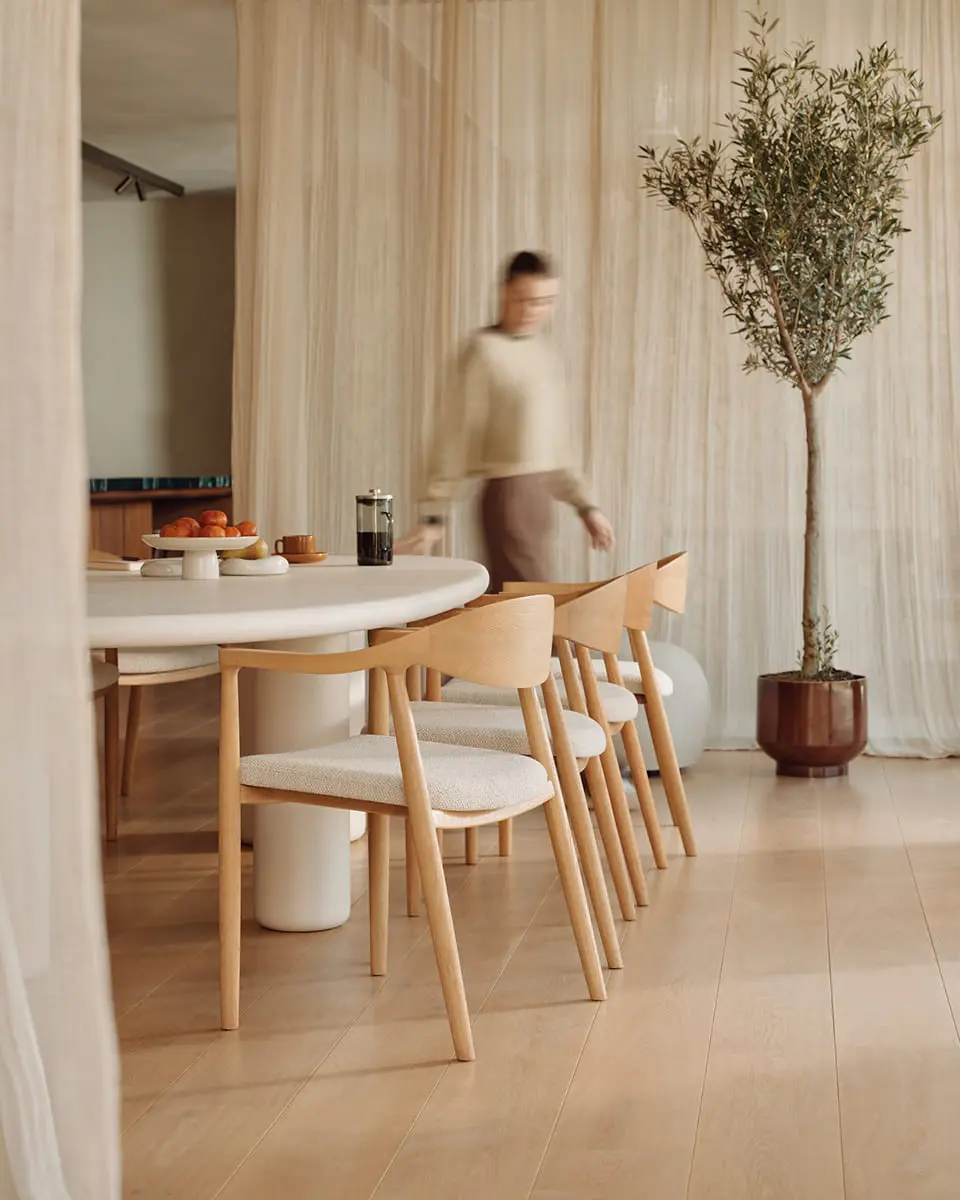 Moderne eetkamer met houten stoelen, ronde tafel en een olijfboom in een lichte, minimalistische ruimte.