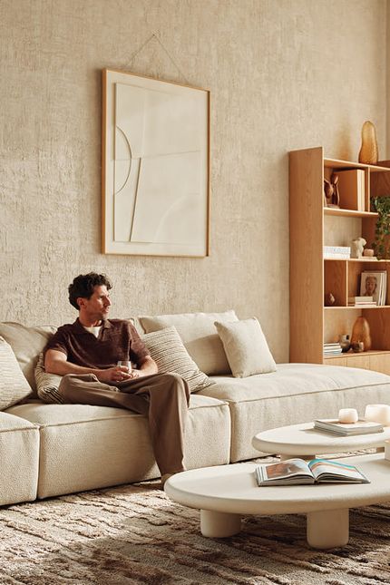 Persoon ontspant op een lichte hoekzetel in een warm interieur met organisch gevormde salontafels en fauteuil.
