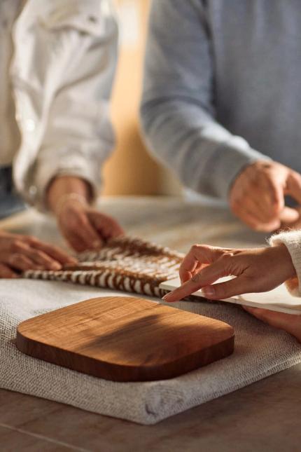 Drie personen bekijken stofstalen op tafel; een hand wijst naar een geweven staal naast een houten sample op een grijze stof.