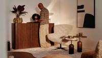 Personne choisissant un disque vinyle près d’un buffet en bois, dans un intérieur chaleureux avec fauteuil clair, tables d’appoint et œuvre murale abstraite.