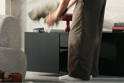Une personne place une couverture roulée dans le tiroir ouvert d'un buffet en bois foncé, avec une lampe de table bordeaux au-dessus.