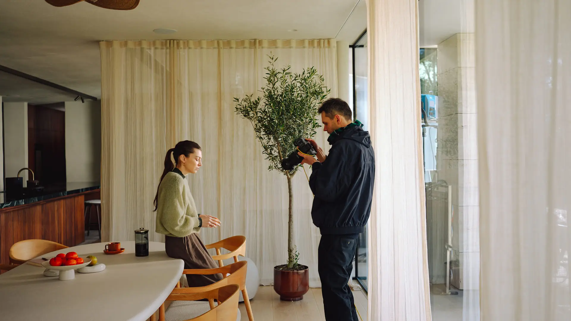 Fotograaf maakt beelden van een vrouw aan de eettafel in een lichte ruimte met houten stoelen, fruit en kamerplant bij het raam.