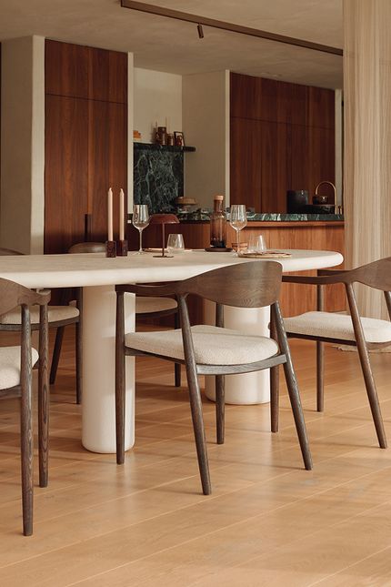 Eettafel met afgeronde hoeken en witte kolompoten, omringd door houten stoelen met lichte zittingen, in een warme eetkamer met houten kasten en keukenblad.