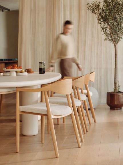 Une femme portant un pull beige marchant autour d'une table ronde avec des chaises en bois. Sur la table, il y a un bol de fruits et une tasse de café, et un olivier se trouve à droite.