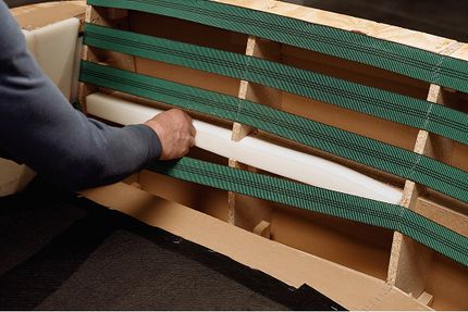 Hand plaatst een witte schuimrol in een houten zetelframe met groene elastische banden.