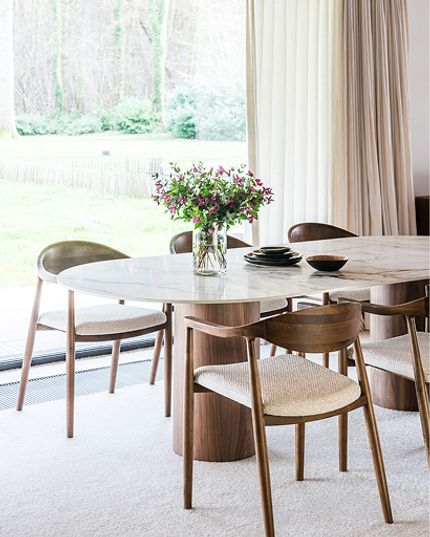 Lichte eetkamer met een afgeronde eettafel met wit marmerlook blad en houten onderstel, omringd door houten stoelen met licht gestoffeerde zittingen.
