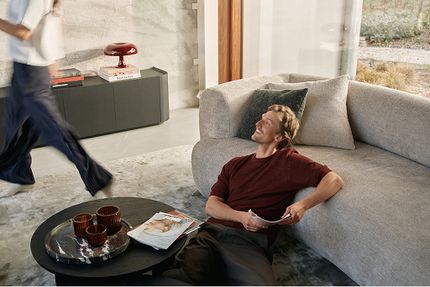 Une personne est assise détendue par terre contre un canapé Tortona clair, avec une table basse noire et des magazines sur un plateau.