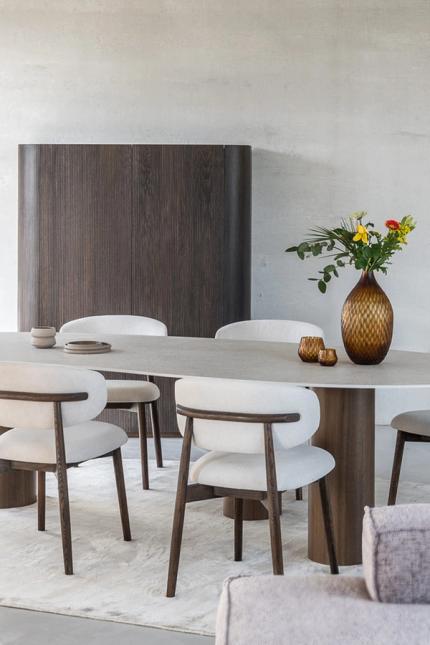 Moderne eethoek met een ovalen tafel, gestoffeerde stoelen en een bloemenvaas op tafel. Op de achtergrond is een houten kast en een persoon zichtbaar die door de kamer loopt.