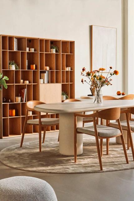 Moderne eetkamer met een ronde tafel en beige stoelen, een houten rek op de achtergrond met planten en boeken, en een vaas met kleurrijke bloemen op de tafel.