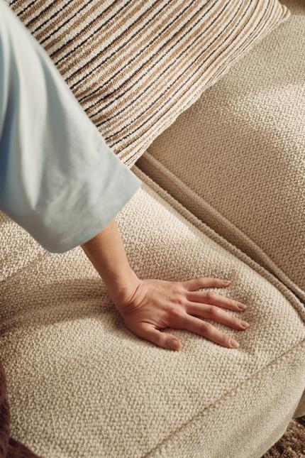 Gros plan d'une main sur l'assise d'un canapé beige clair en tissu texturé, avec un coussin rayé sous une lumière douce.