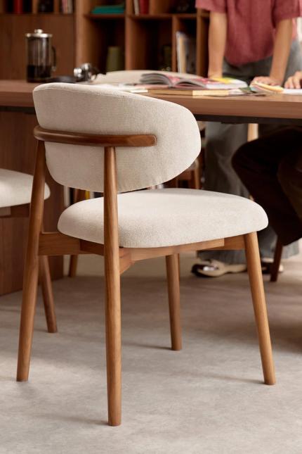 Close-up van een eetkamerstoel met licht gestoffeerde zitting en rug, op een warm houten frame; op de achtergrond een tafel met mensen en boeken.
