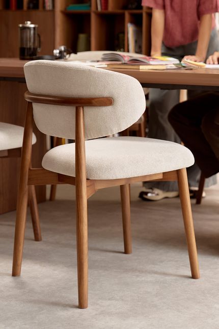 Close-up van een eetkamerstoel met licht gestoffeerde zitting en rug, op een warm houten frame; op de achtergrond een tafel met mensen en boeken.