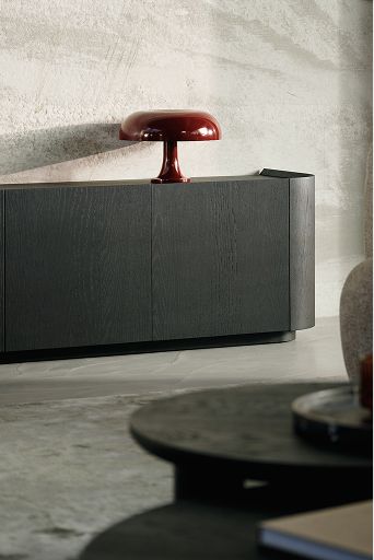 Gros plan d'un buffet en bois foncé contre un mur en béton, avec une pile de livres et une lampe de table bordeaux ; au premier plan, une table basse ronde floue.