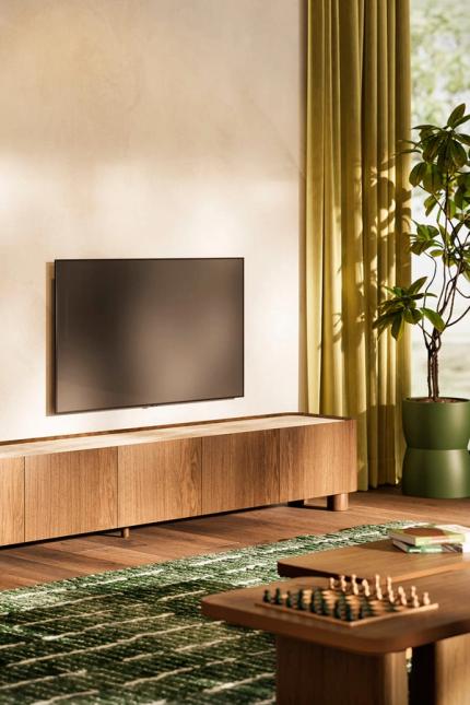 Meuble TV long en bois avec coins arrondis sous une télévision murale, décoré d'un vase vert et baigné de la lumière chaude du soleil.