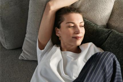 Femme allongée de manière détendue sur un canapé en tissu gris avec des coussins, le bras au-dessus de la tête, baignée dans une lumière chaude du soleil.