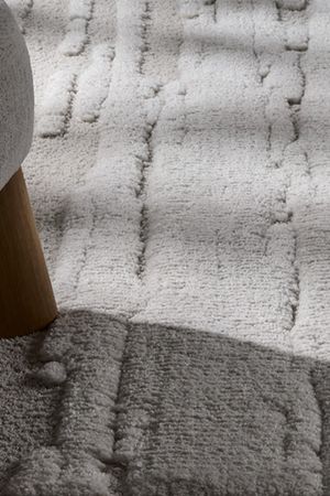 Detail van een licht tapijt met reliëfpatroon naast de houten poot van een fauteuil.