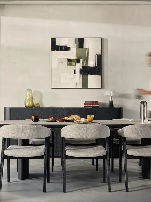 Longue table à manger avec quatre chaises grises et pieds foncés, nature morte sur la table, tableau abstrait au mur et personne en mouvement.