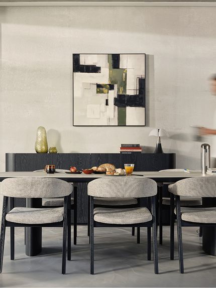 Longue table à manger avec quatre chaises grises et pieds foncés, nature morte sur la table, tableau abstrait au mur et personne en mouvement.