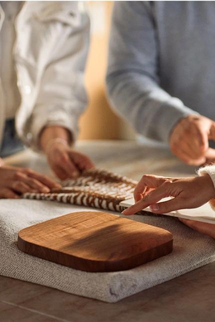 Trois mains de personnes touchant ensemble un tissu tissé, avec une planche à découper en bois sur un tissu en lin sur la table. Les mains sont dans différentes positions, manipulant le matériau.