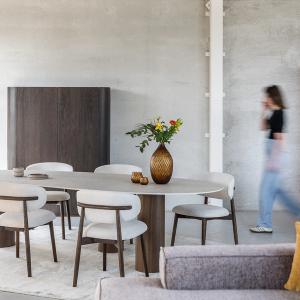 Moderne eetkamer met een ovale tafel, houten stoelen met beige kussens, een vaas met bloemen op de tafel, en een kast op de achtergrond. Een persoon beweegt zich door de ruimte.
