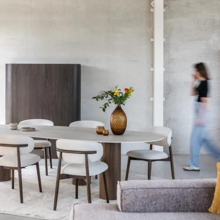Moderne eetkamer met een ovale tafel, houten stoelen met beige kussens, een vaas met bloemen op de tafel, en een kast op de achtergrond. Een persoon beweegt zich door de ruimte.