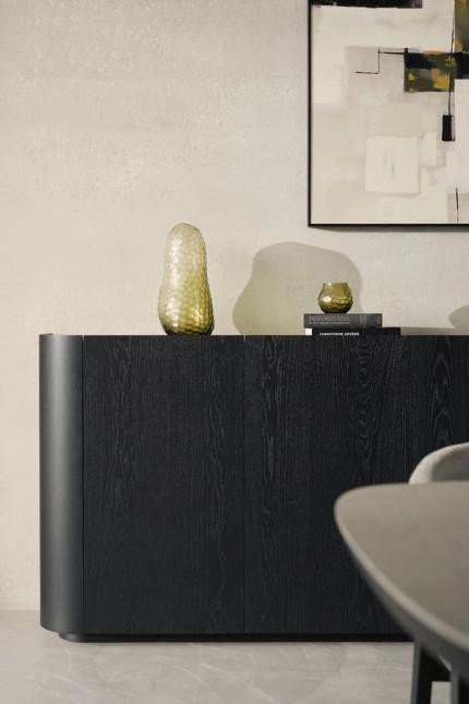 Gros plan d'un buffet en bois foncé avec un côté arrondi, décoré d'un vase et de livres contre un mur clair et une œuvre d'art abstraite.