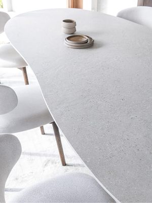 Detailbeeld van een lichte eettafel met een zacht afgerond, organisch gevormd tafelblad en subtiele steenstructuur, omringd door licht gestoffeerde stoelen met houten poten.