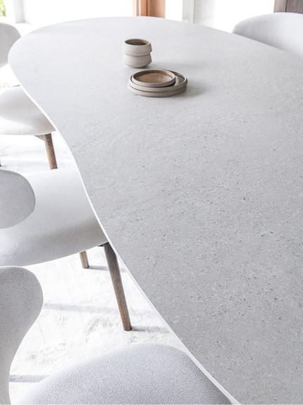 Detailbeeld van een lichte eettafel met een zacht afgerond, organisch gevormd tafelblad en subtiele steenstructuur, omringd door licht gestoffeerde stoelen met houten poten.