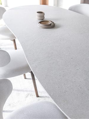 Detailbeeld van een lichte eettafel met een zacht afgerond, organisch gevormd tafelblad en subtiele steenstructuur, omringd door licht gestoffeerde stoelen met houten poten.