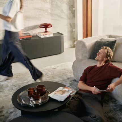 Een man die ontspannen op een comfortabele bank ligt, met een glimlach op zijn gezicht. In de achtergrond beweegt een vrouw langs een stijlvolle zwarte kast, met op de salontafel tijdschriften en glazen vazen. Het lichte interieur straalt rust en gezelligheid uit.