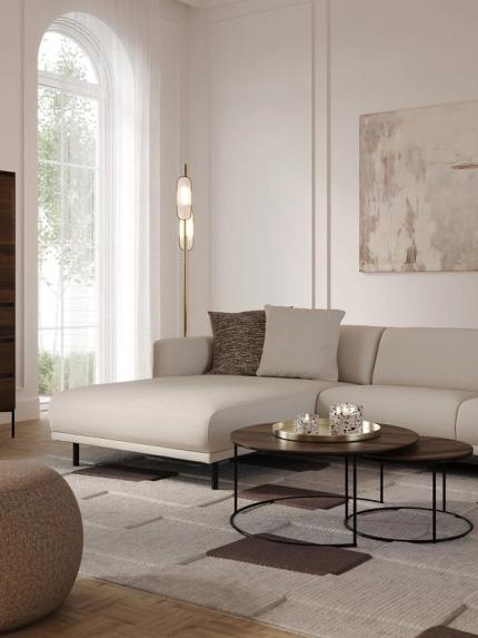 Lichte, compacte zithoek met chaise longue, twee ronde salontafels met houten blad en vloerlamp naast een groot boograam.
