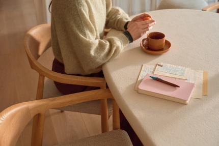 Personne assise sur une chaise en bois à une table ovale, tasse de café, carnet et stylo; tient une mandarine.