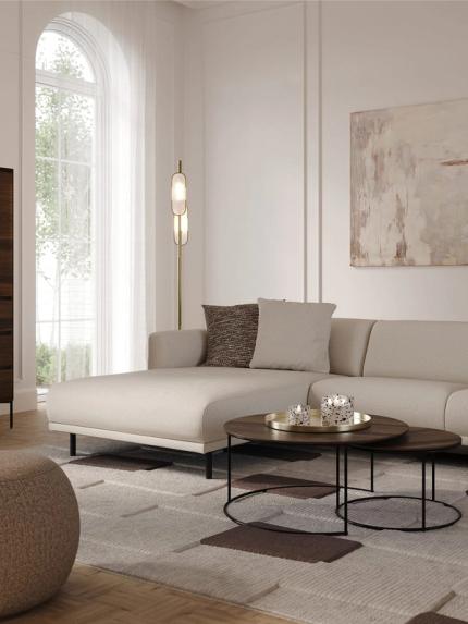 Canapé d’angle Evora beige clair avec méridienne dans un salon lumineux, avec tables basses rondes, lampadaire et tableau abstrait.