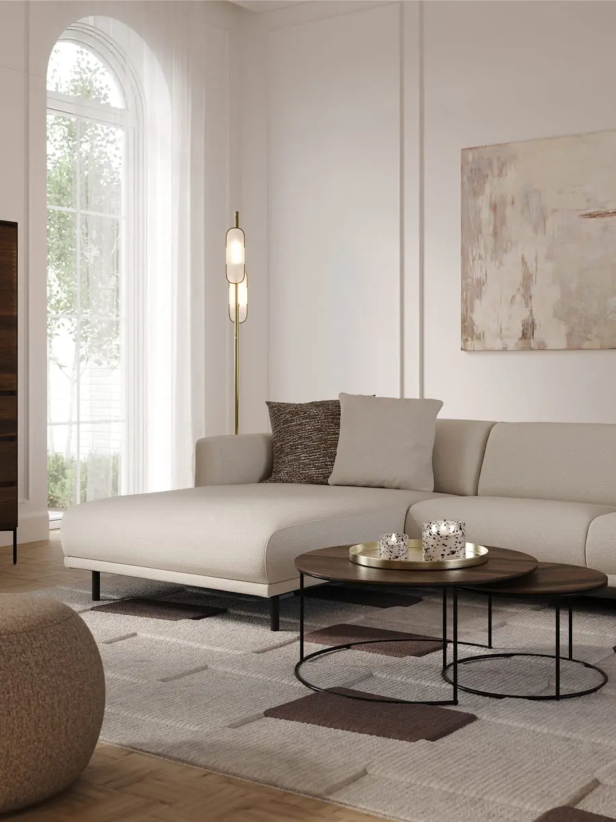 Canapé d’angle Evora beige clair avec méridienne dans un salon lumineux, avec tables basses rondes, lampadaire et tableau abstrait.