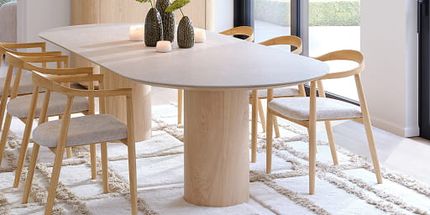 Lichte eetkamer met een grote eettafel met afgeronde hoeken en een licht blad op cilindervormige houten poten, omringd door houten armstoelen met licht gestoffeerde zittingen.