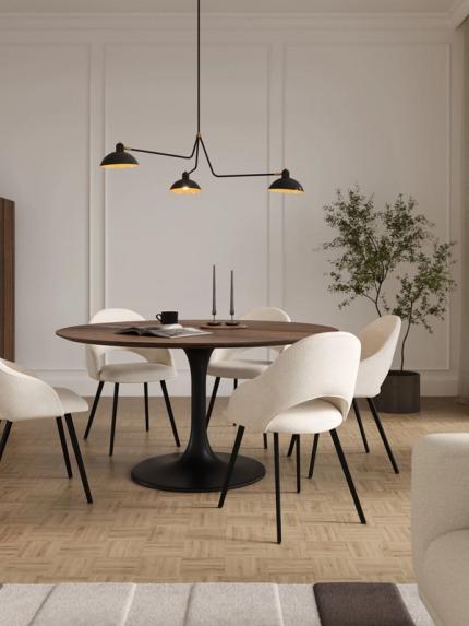 Salle à manger lumineuse avec table ronde foncée sur pied central, quatre chaises beige, suspension noire et grand meuble en bois près de grandes fenêtres.