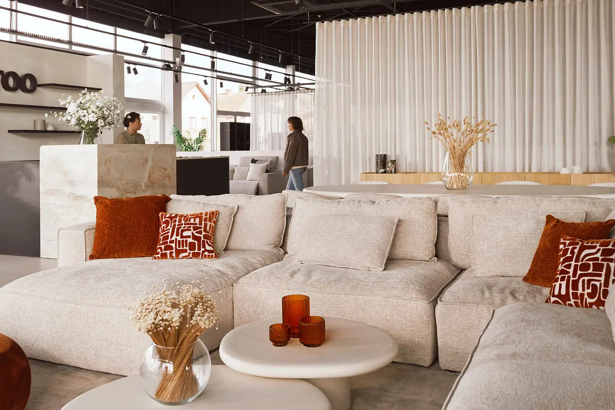 Showroom met lichte banken, oranjerode kussens en decoratieve objecten, met een persoon die door de ruimte beweegt.