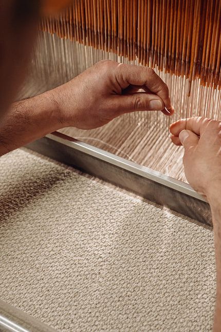 Gros plan d’un artisan guidant les fils à la main sur un métier à tisser, tandis qu’un textile clair à la texture fine prend forme.