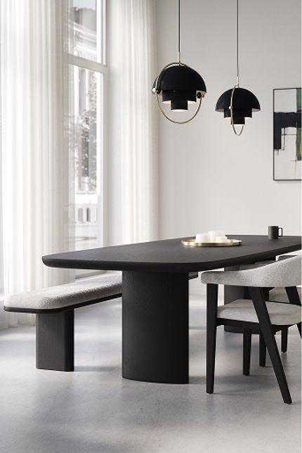 Lichte eetkamer met zwarte eettafel, stoelen en bankje, hanglampen, hoge ramen met witte gordijnen en grote kamerplant.
