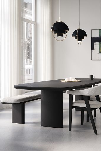 Lichte eetkamer met zwarte eettafel, stoelen en bankje, hanglampen, hoge ramen met witte gordijnen en grote kamerplant.